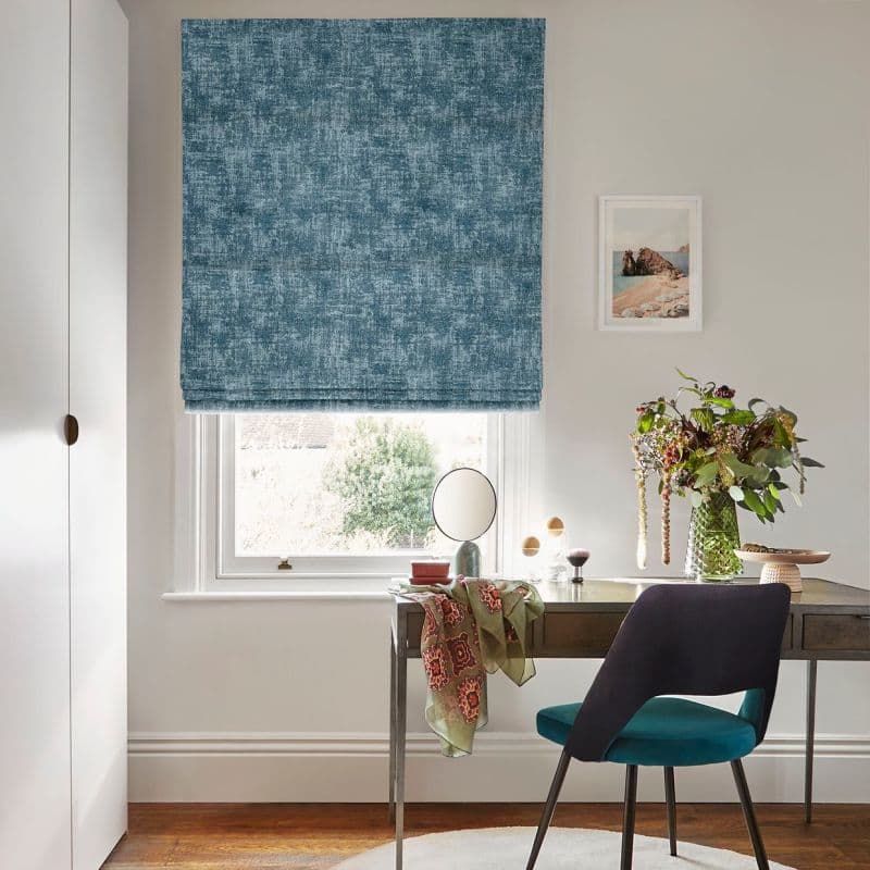 Blue textured Roman blind over the window, folded and partially lowered, filtering daylight; compact desk/vanity with teal chair, round mirror, vase of flowers, pale walls, framed seaside print, garden view.