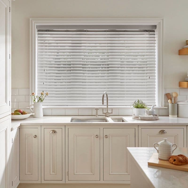 White horizontal Venetian blinds fully covering a large window, slats slightly tilted to filter soft daylight; above a cream kitchen sink and marble countertop with faucet, plants, and utensils.