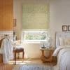 Floral-patterned Roman blind, mostly lowered, filtering soft daylight through a sash window; cozy sunlit bedroom with white bed, wicker bedside table, dressing table and leafy outside view.
