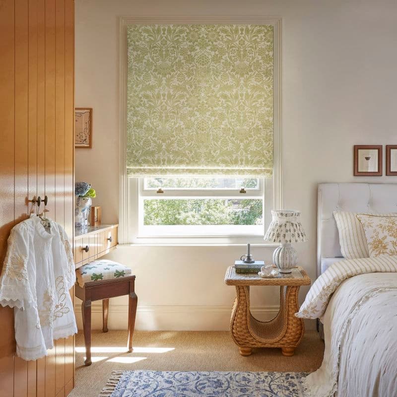 Floral-patterned Roman blind, mostly lowered, filtering soft daylight through a sash window; cozy sunlit bedroom with white bed, wicker bedside table, dressing table and leafy outside view.