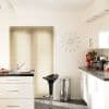 Beige horizontal Venetian blinds covering a central window, closed and softly filtering daylight; modern white kitchen with dark countertops, a black barstool, stainless bin, wall clock and tulip bouquet.