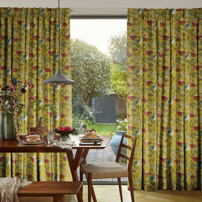Floral pleated full-length curtains (mustard yellow with red and blue flowers) pulled aside, framing a glass sliding door; dining table with breakfast setting under a pendant lamp, sunlit garden beyond.