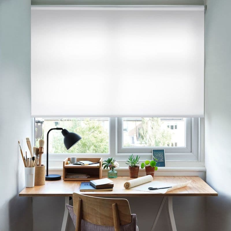 White roller blind lowered across the upper window, smooth translucent fabric with visible roller; filtering soft daylight onto a wooden desk with a lamp, brushes, notebooks and small potted plants, trees outside.