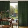 Thick dark-green pleated curtains, floor-to-ceiling and drawn aside to frame a central opening; partially open, letting daylight onto a wooden dining table set for breakfast and sunlit garden trees.