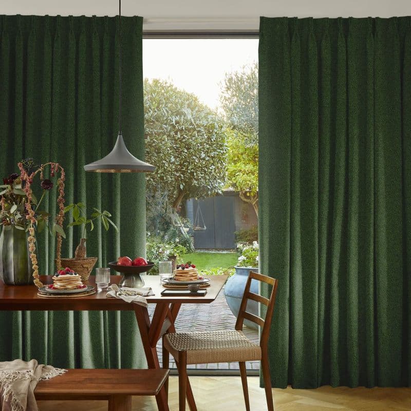 Thick dark-green pleated curtains, floor-to-ceiling and drawn aside to frame a central opening; partially open, letting daylight onto a wooden dining table set for breakfast and sunlit garden trees.