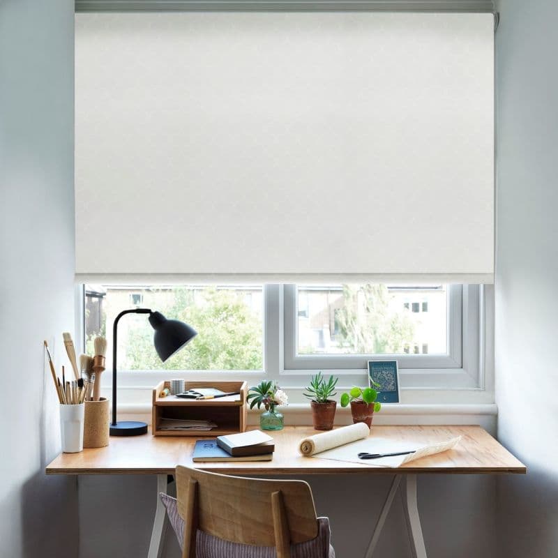 White patterned roller blind, pulled down covering most of the window and softly diffusing daylight; small home workspace with wooden desk, black lamp, potted plants and stationery below.