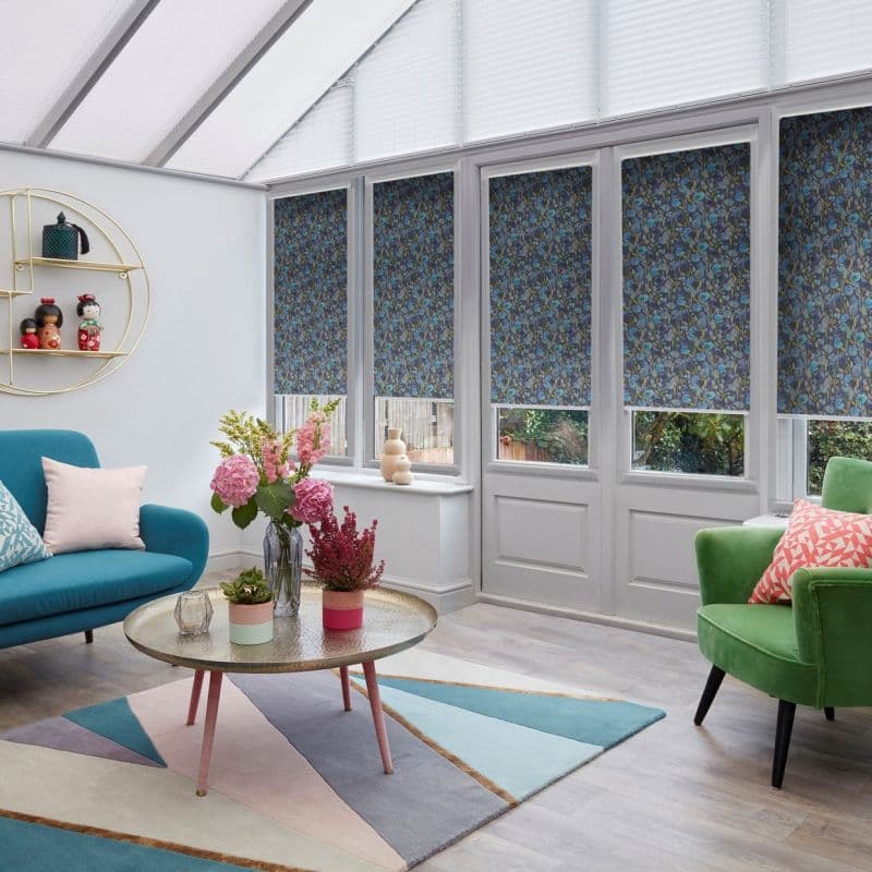 Blue floral roller blinds lowered over tall conservatory windows and doors, filtering daylight; bright sunroom furnished with teal sofa, green armchair, coffee table with vases and pastel geometric rug.