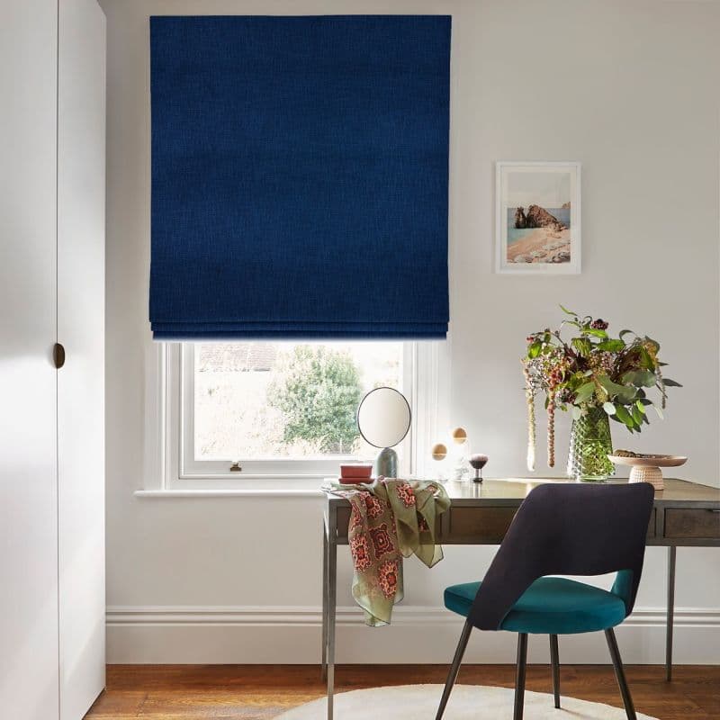 A navy-blue Roman blind, lowered over the window with soft folds at the base, blocking most light; set above a desk in a bright, minimalist home office with vase and chair.