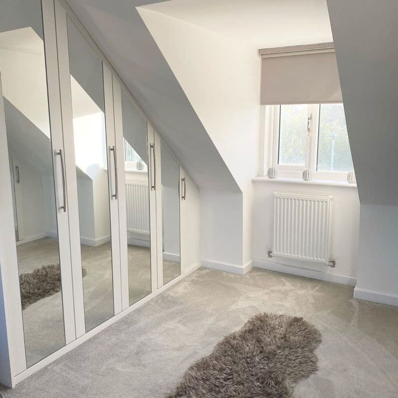 A beige roller blind partially lowered over a dormer window, filtering daylight; mirrored wardrobe doors reflect the angled white attic room with radiator, pale carpet, and a gray shag rug.