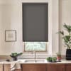 Dark gray roller blind covers a kitchen window above a stainless sink; sunlight filters below, revealing leafy trees. Marble countertop holds bowls of greens, potted plants and framed sketch.