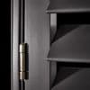 Matte dark louvered window shutters with angled horizontal slats and a brass hinge at left — slats closed, casting sharp shadows in a close-up interior view with low directional lighting.