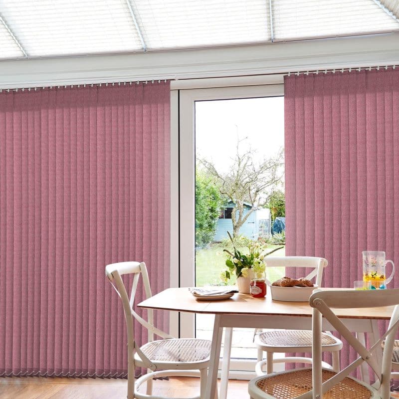 Vertical pink fabric blinds, mostly closed with a central gap, filtering bright daylight; wooden dining table with cane‑seat chairs inside, framed view of a tree and garden shed outside.