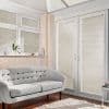 Pleated cream blinds fully lowered on casement windows and French doors, filtering sunlight and casting striped shadows across a living room with tufted sofa, hardwood floor, rug, and plant.