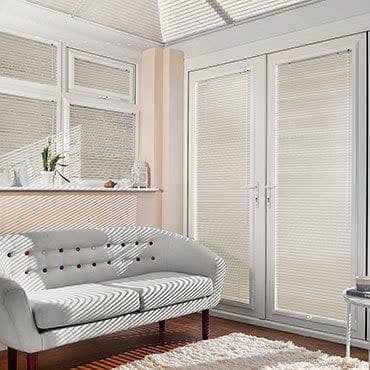 Pleated cream blinds fully lowered on casement windows and French doors, filtering sunlight and casting striped shadows across a living room with tufted sofa, hardwood floor, rug, and plant.