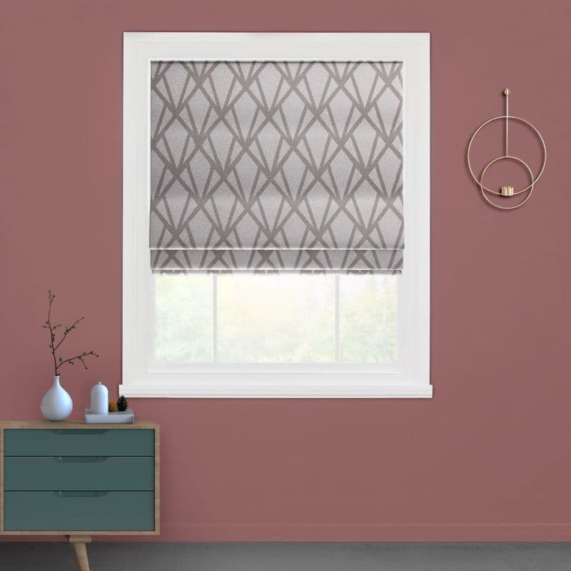 A grey geometric-patterned Roman shade lowered over a white-framed window, diffusing daylight; set in a mauve-walled room with a teal drawer, small vases on a console, and circular gold wall decor.