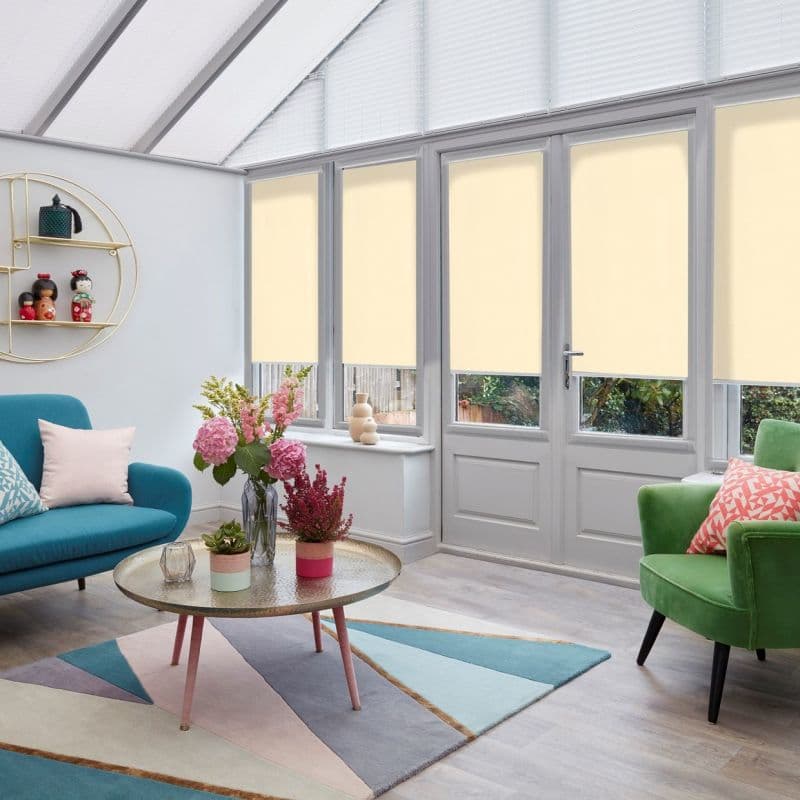 Cream roller blinds lowered over four tall conservatory windows and door, filtering daylight while leaving lower panes clear; bright living room with sofas, rug, coffee table and garden view.