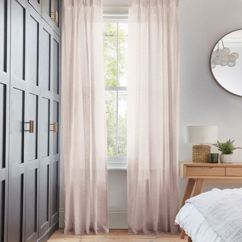 Sheer pale-blush, floor-length gathered curtains framing a tall sash window, filtering soft daylight through semi-sheer fabric; bedroom context with wooden nightstand, lamp, round mirror, and dark paneled wardrobe.