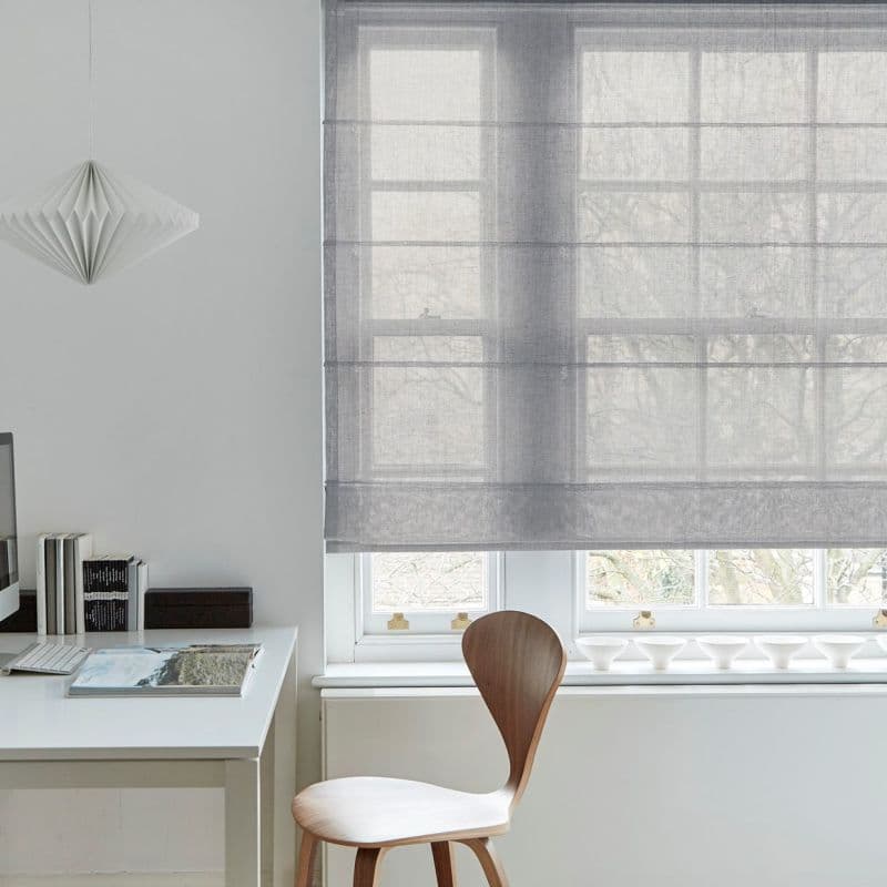 Sheer grey Roman blind, lowered with horizontal ribs, filtering soft daylight and faint tree silhouettes; in a minimalist home office with white desk, wooden chair and a paper pendant.
