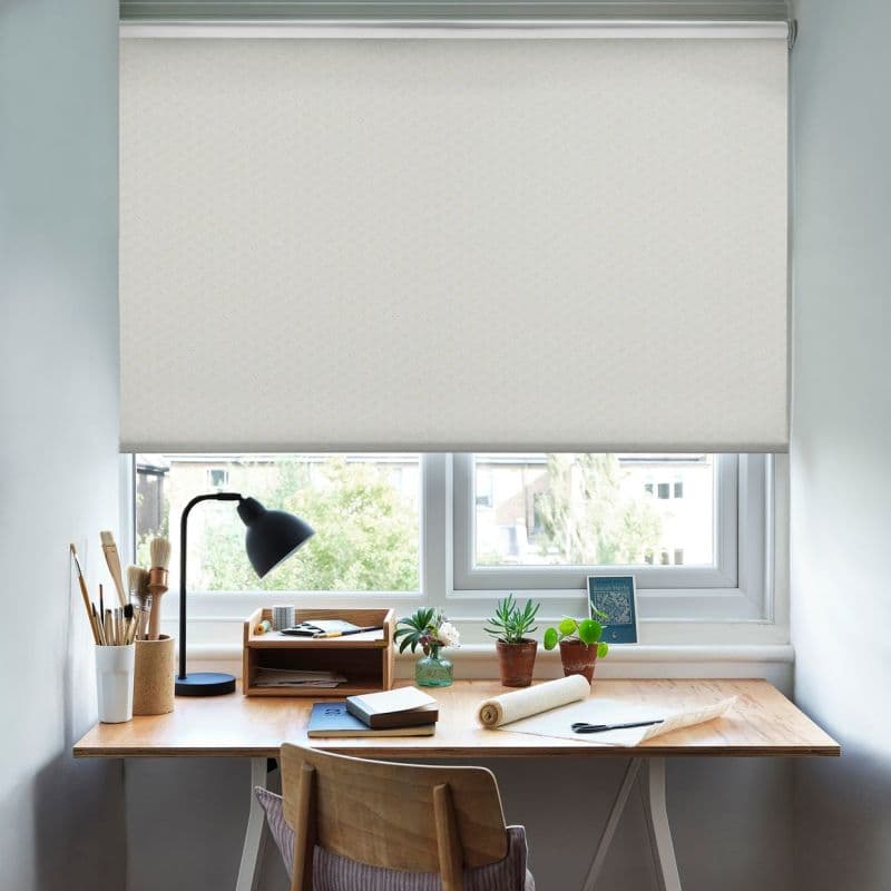 A white patterned roller blind pulled down mid-window, softly filtering daylight; below, a wooden desk with lamp, brushes, notebooks and small potted plants in a bright, tidy home workspace.