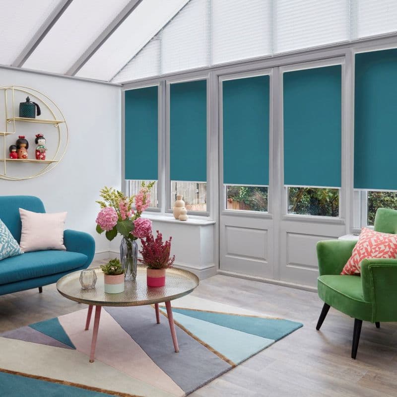 Teal roller blinds on four tall conservatory windows/doors, lowered across upper panes leaving narrow lower glass visible; closed to reduce light and view in a sunroom with chairs and flowers.