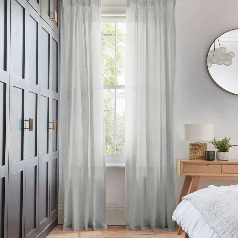 Sheer floor-length curtains, light gray and pleated, hung at a tall sash window; they filter soft daylight and are parted, in a bedroom with dark paneled wardrobe and wooden nightstand.
