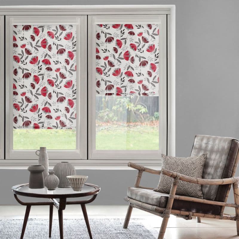 Floral roller blinds (white with red and gray flowers) covering two windows—left fully lowered, right halfway—filtering daylight into a gray-walled living room with chair and coffee table; garden visible.