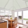 White pleated cellular blinds cover the pitched conservatory ceiling and all windows and doors, closed and softly diffusing daylight, in a bright kitchen-dining area with wooden table and garden view.