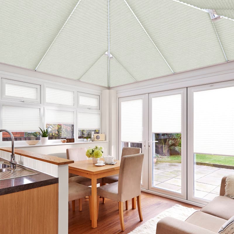 White pleated cellular blinds cover the pitched conservatory ceiling and all windows and doors, closed and softly diffusing daylight, in a bright kitchen-dining area with wooden table and garden view.