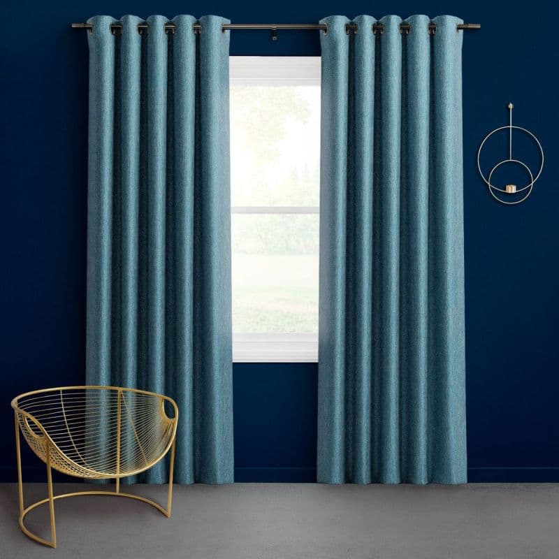 Teal grommet-top curtains, full-length panels on a metal rod, drawn slightly apart and filtering light from a window; navy-painted room with gray carpet, brass wire chair and circular wall sconce.