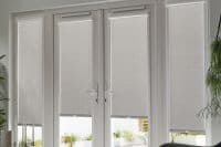 Grey roller blinds with top cassette housings across four tall window/door panels, lowered to filter light with narrow bottom gaps; central French doors, potted plants in a bright modern room.