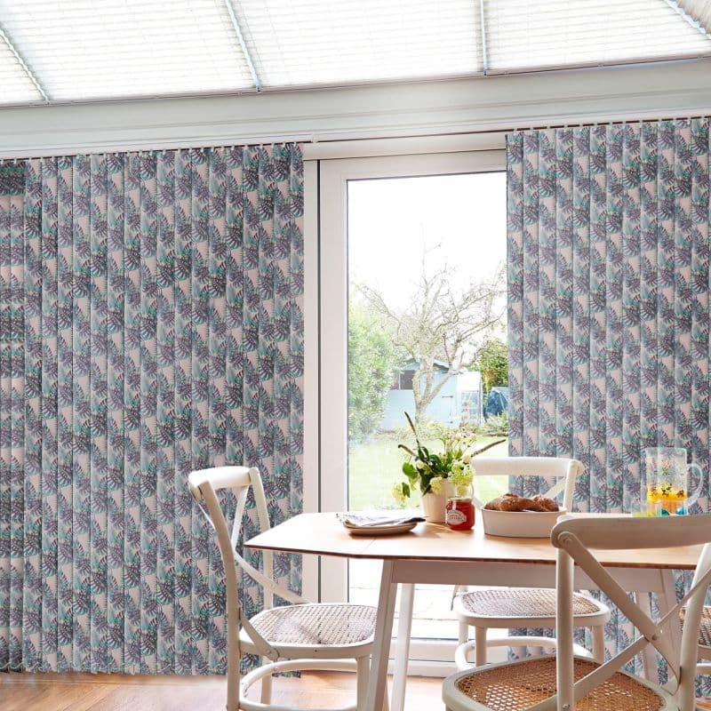 Patterned vertical blinds covering a sliding glass door, partially drawn to reveal a central gap and filter daylight; small dining table with chairs, vase and bowl, view of garden and shed beyond.