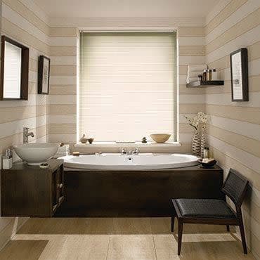 A light-filtering pleated cellular blind fully lowered over the window, softly diffusing daylight into a contemporary striped-wall bathroom with a central soaking tub, wood vanity, mirror, shelf, and chair.