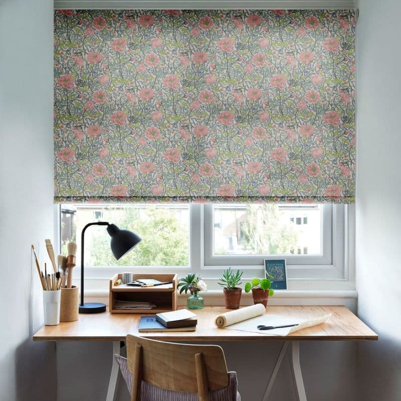 Floral roller blind with pink poppies lowered across the window, filtering daylight — above a wooden desk holding a lamp, brushes, small potted plants and stationery in a bright workspace.