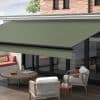 Green retractable fabric awning, fully extended with black front bar, shading ground-floor glass patio doors; outdoor terrace with wicker armchairs, round coffee table, cushioned bench and brick façade in daylight.