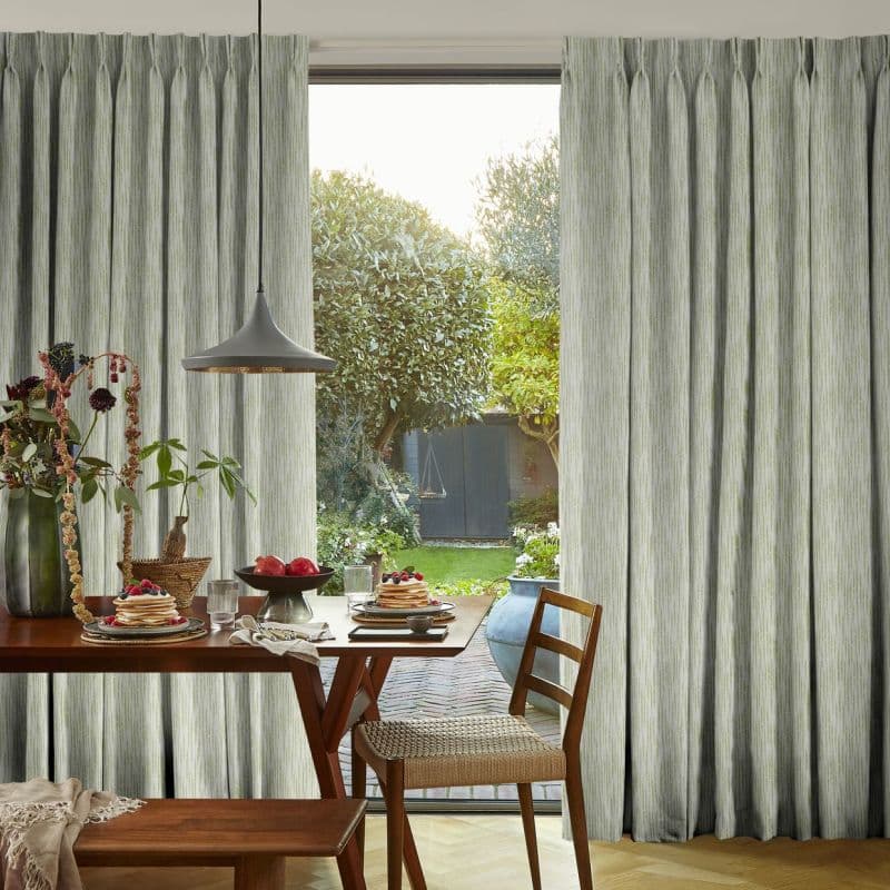 Full-length pale green textured pinch-pleat curtains, parted at center, framing and softening daylight; beyond a sunlit garden, foreground wooden dining table set with pancakes, vase, pendant lamp and chairs.