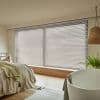 White horizontal Venetian blinds covering a wide ground-level window, slats mostly closed and gently filtering soft daylight, in a warm, minimalist bedroom with wood floor, rattan accents, and a potted fern.