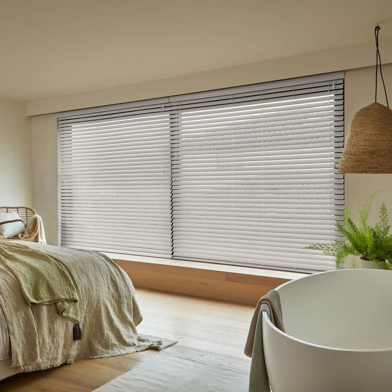 White horizontal Venetian blinds covering a wide ground-level window, slats mostly closed and gently filtering soft daylight, in a warm, minimalist bedroom with wood floor, rattan accents, and a potted fern.