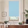 Light-blue roller blind partially lowered over a window above a kitchen sink; countertop holds faucet, bowls of leafy vegetables, a potted plant, and a framed sketch on the wall.