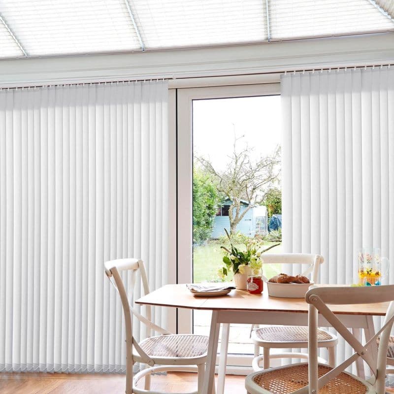 White vertical blinds, full-length and mostly closed with central gap, filtering daylight and parted to reveal a glass sliding door; dining table and chairs in a bright garden-facing room.