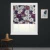 Floral Roman blind, folded and partially lowered, filtering soft daylight; in a white window frame on a dark charcoal wall, beside a wooden side table with glass-domed lamp and abstract print.