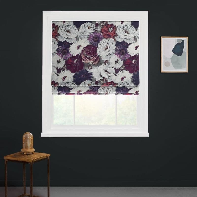 Floral Roman blind, folded and partially lowered, filtering soft daylight; in a white window frame on a dark charcoal wall, beside a wooden side table with glass-domed lamp and abstract print.