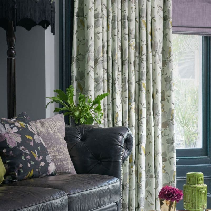 Full-length pleated floral curtains (pale green with grey-yellow blooms), drawn closed, softly filtering daylight; living room setting with black tufted leather sofa, cushions, potted fern, and a teal window frame.