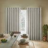 Grommet-top gray curtains, hanging on a metal rod, drawn into pleats with a narrow central gap, filtering daylight in a sunlit dining room with wooden table and potted plant.
