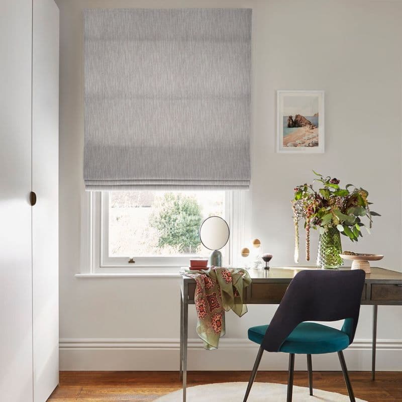 Grey textured Roman blind, lowered and slightly folded at bottom, filtering soft daylight through the window; a desk beside it with chair, mirror, vase of flowers and framed seaside print.