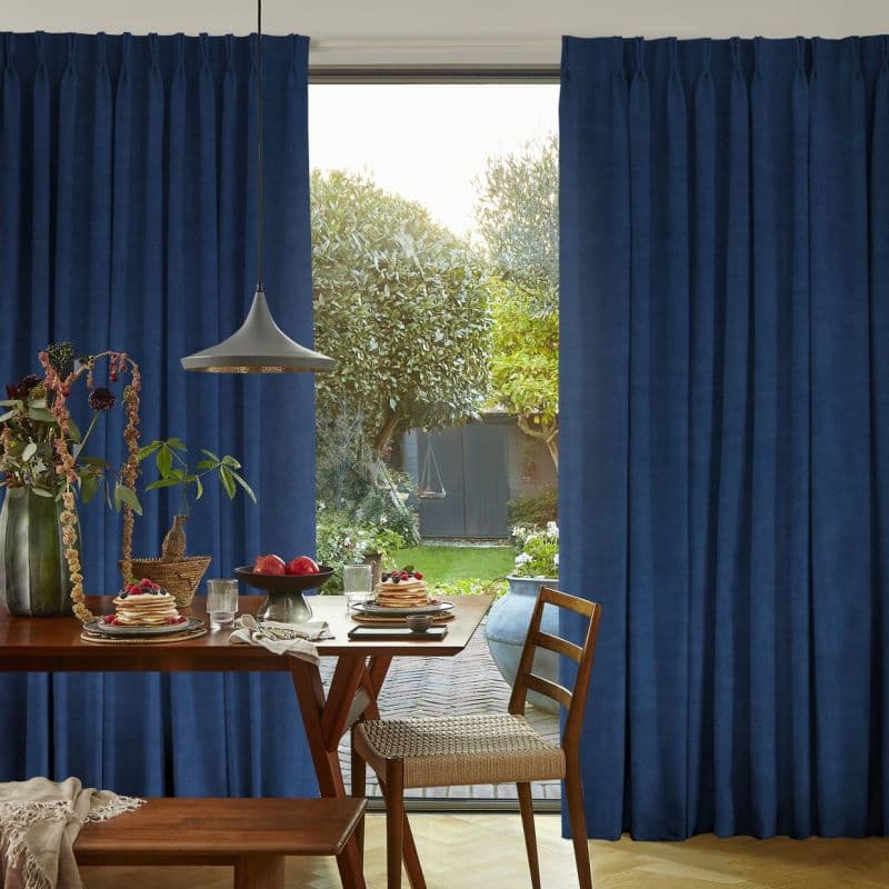 Floor-to-ceiling navy pleated curtains, parted at center, framing a sliding glass door; filtering soft daylight into a dining area where a set table faces a sunlit garden with trees and a shed.