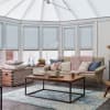 Pale-blue pleated blinds fitted inside bay windows and the conservatory roof, lowered and gently diffusing daylight; cozy sunroom living area with sofa, chunky pink throw, wooden coffee table, blue rug.