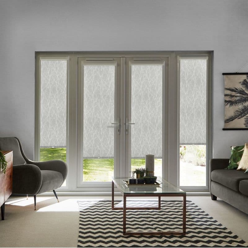 White roller blinds with a subtle branch pattern covering four-panel French doors and side windows, lowered and filtering daylight (small bottom gaps reveal lawn) — modern living room, soft natural light.
