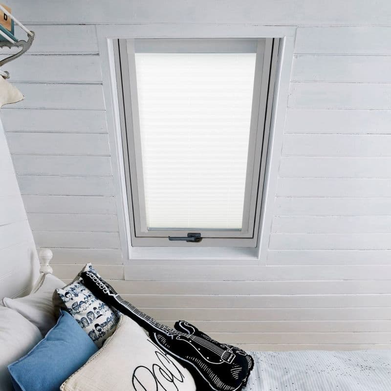 White pleated blind fully lowered over a skylight window, diffusing daylight; set in a white-painted slatted attic ceiling above bed with blue and patterned pillows and a guitar-shaped cushion.