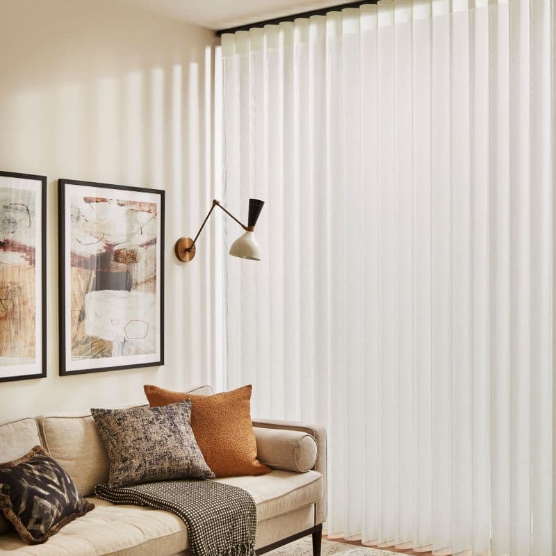 Sheer white vertical curtains, pleated, cover a window/door on the right, drawn closed and diffusing sunlight; a beige sofa with patterned pillows and framed art fills the warm living room.