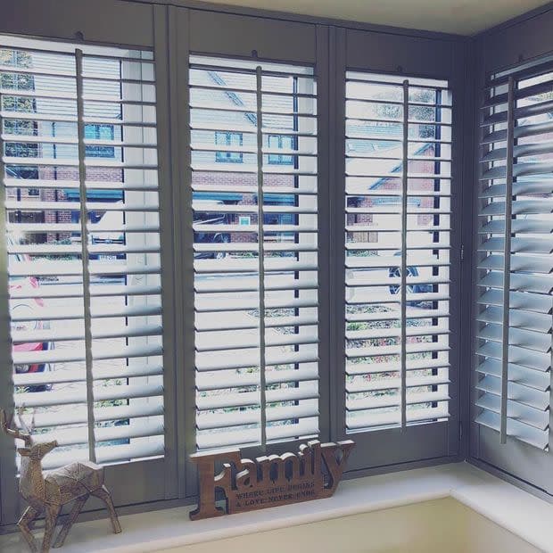 White wooden plantation shutters cover a three-panel bay window, slats tilted open to filter bright daylight and show a suburban street; windowsill holds a wooden "Family" sign and deer ornament.
Transcription:
Family
WHERE LIFE BEGINS & LOVE NEVER ENDS
